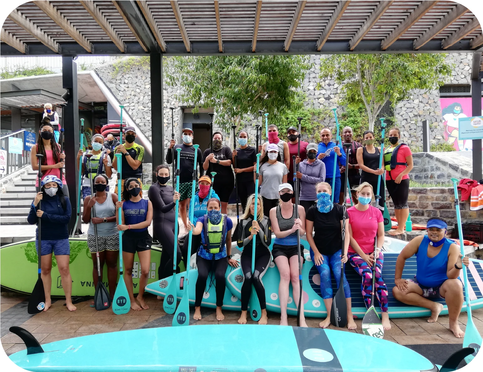 A large group of people gathered on the SUP Cape Town deck holding paddles and looking at the camera. Many of them are wearing medical face masks.