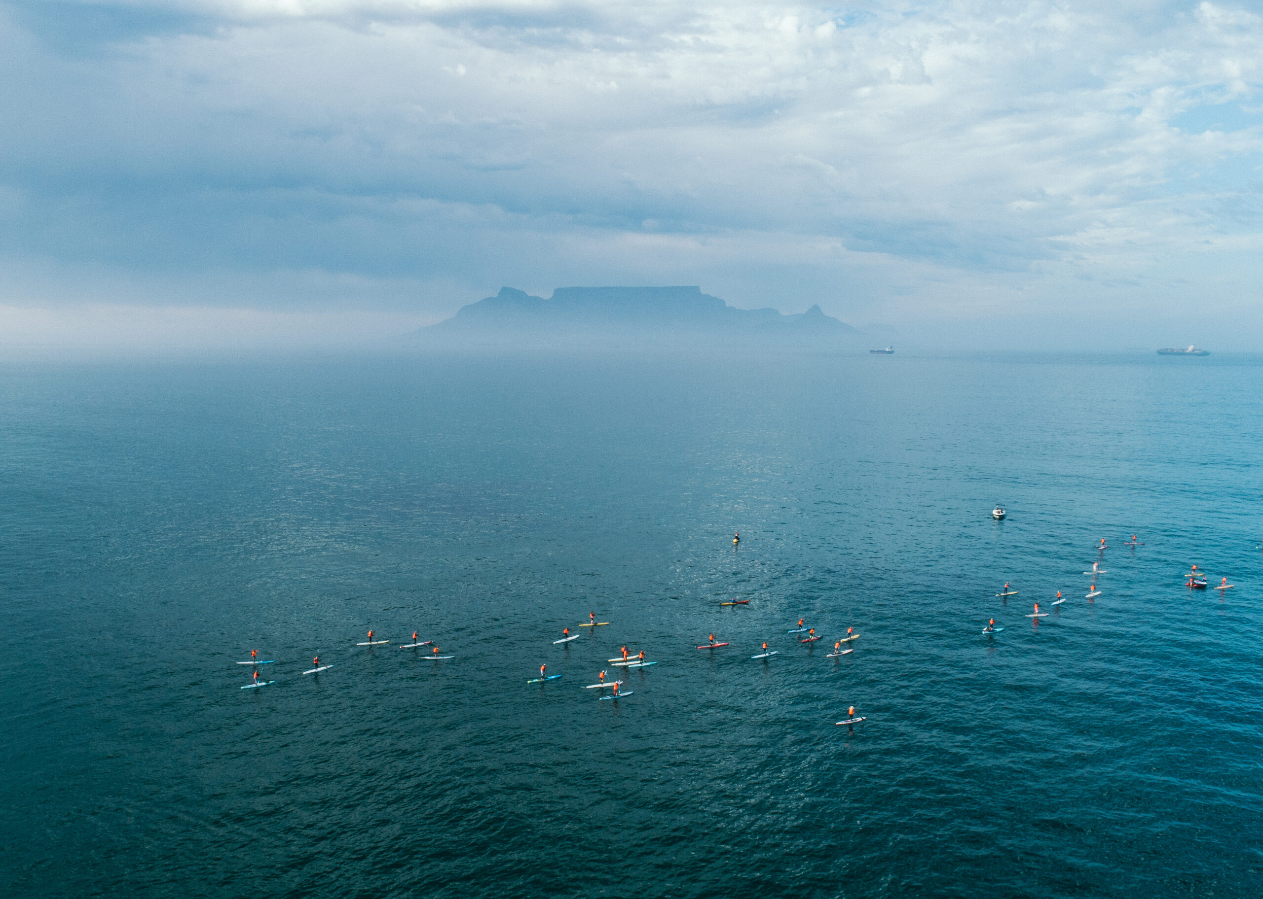 A drone shot showing a large group of people on SUP boards. The outline of Table Mountain is silhouetted through mist in the background.