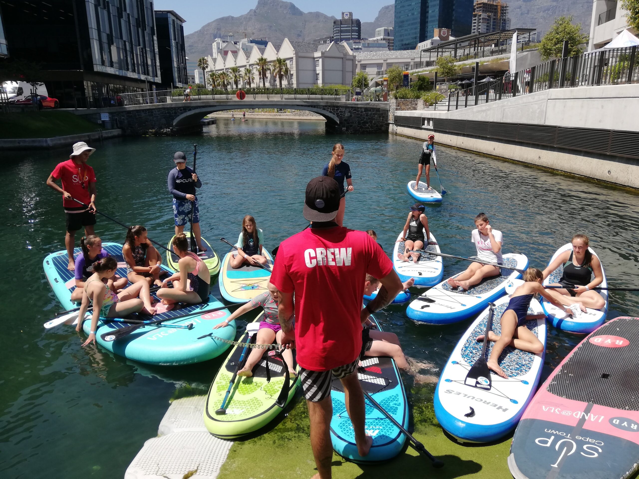 A group of teenagers laying or sitting on sup boards surround a SUP Cape Town Crew member standing on the deck.