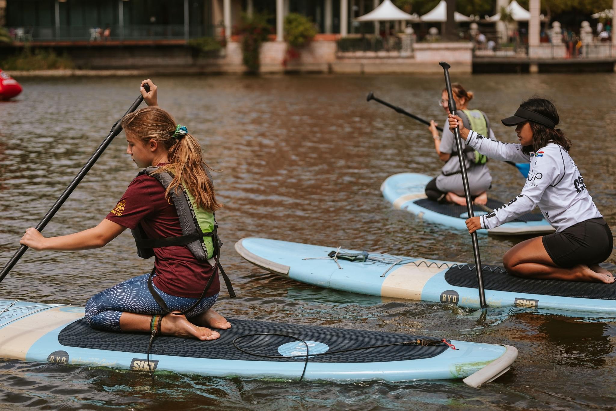 A SUP Cape Town crew member wearing a white shirt instructs two girls paddling on the Intaka canals, they are all kneeling on their boards.
