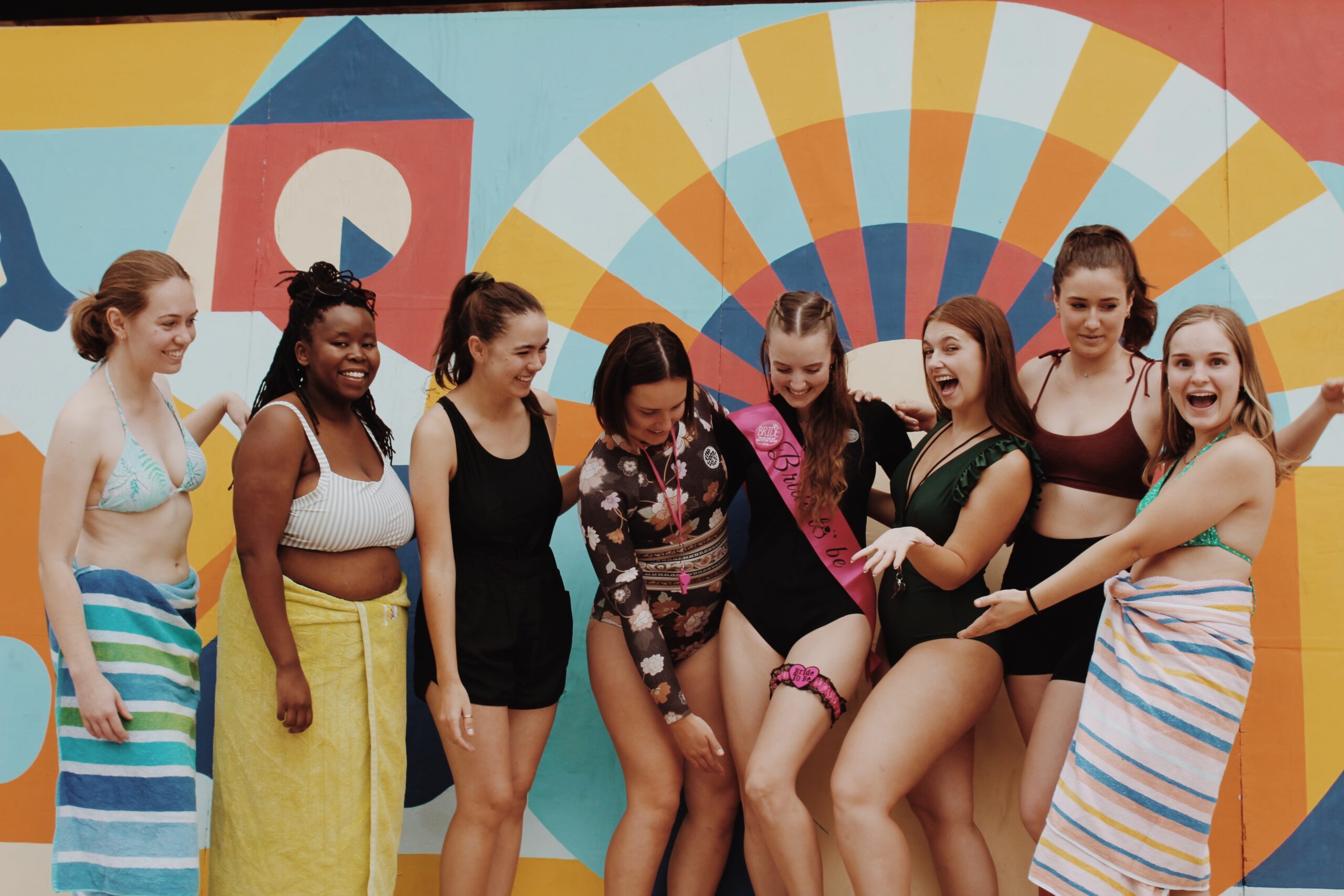 A group of 8 women in bathing suits, centred around a bachelorette wearing a pink 'bride to be' sash.