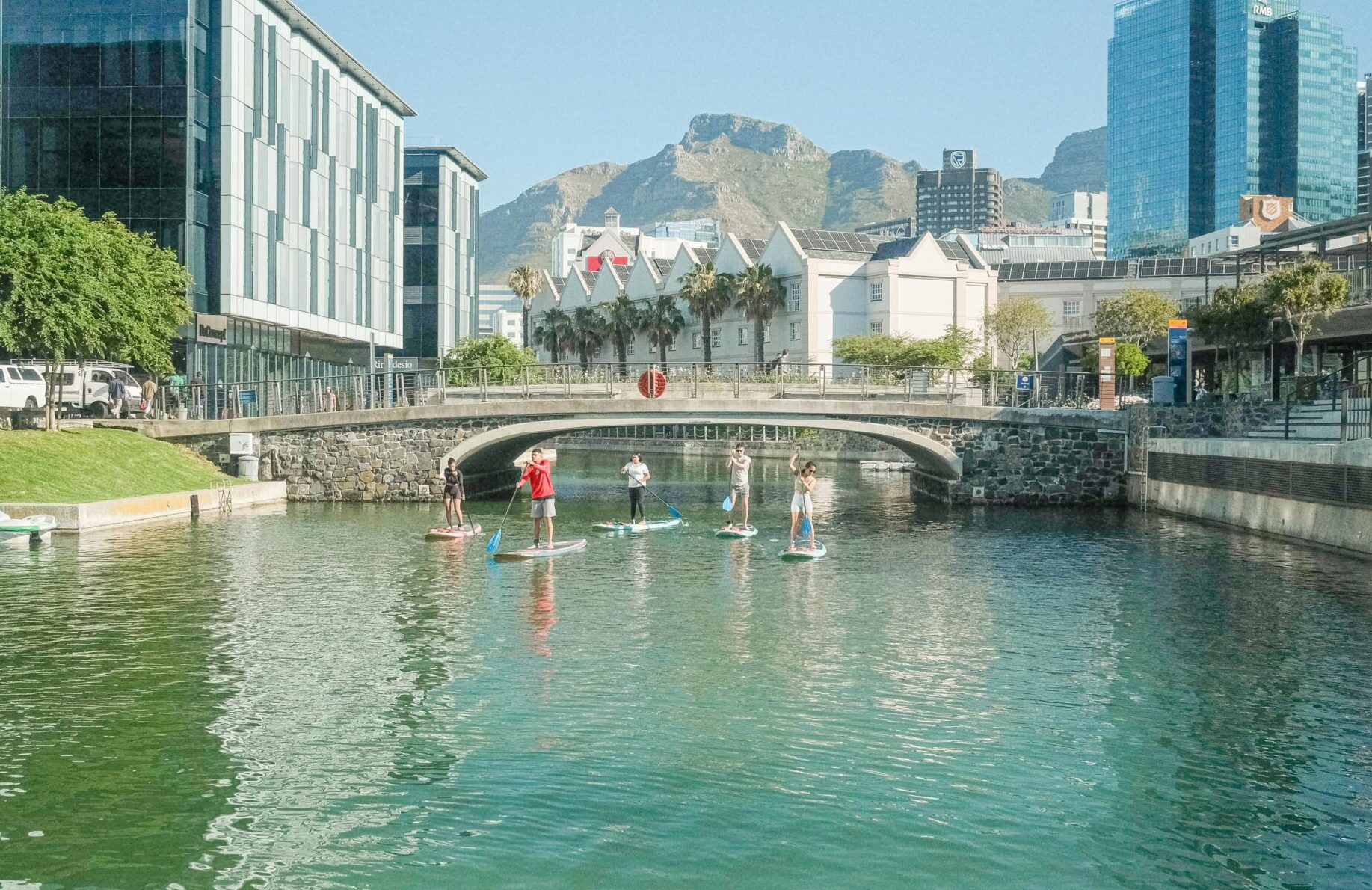 A landscape view of the canal, five people including a SUP instructor are paddling out from under the bridge. Buildings and mountains are in the background.