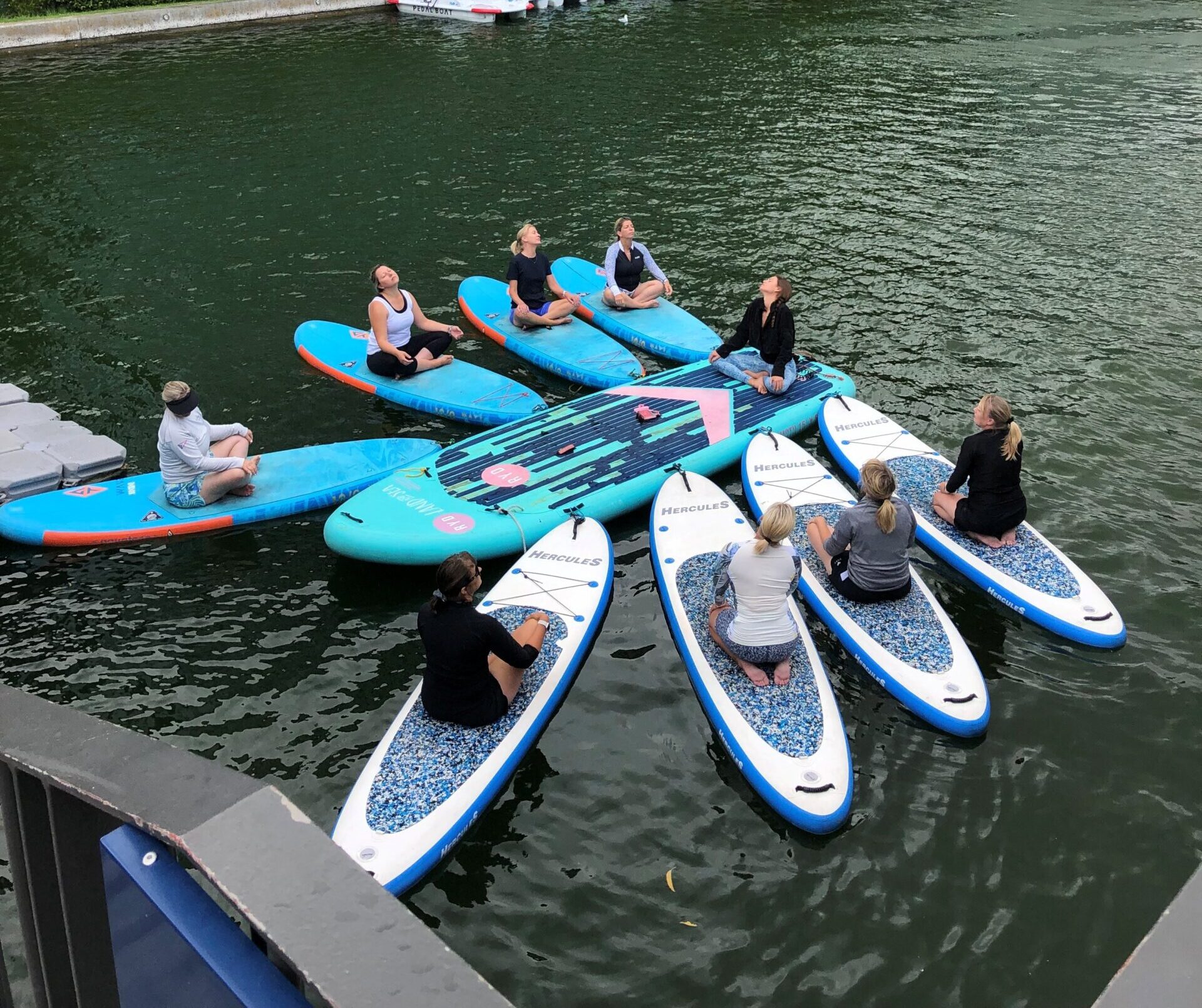 A group of 8 SUPs clustered around one larger board. All the people on them are sat either crossed legged or kneeling.