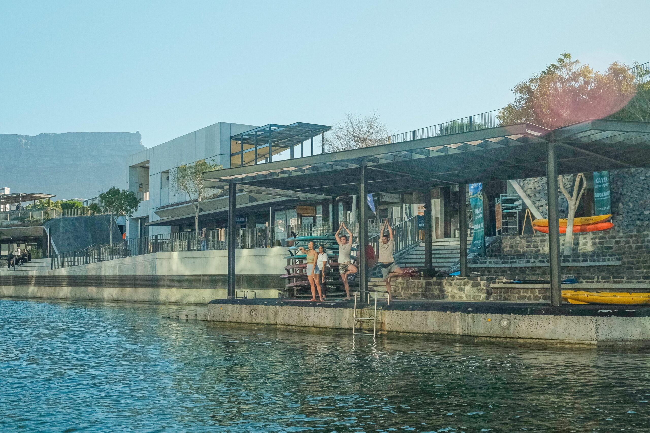 A group of four people stand on yoga mats on the SUP Cape town deck. The photograph is taken from the opposite side of the canal looking over at the deck.